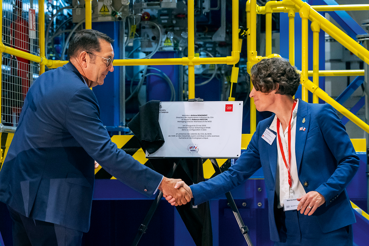Deux personnes en costume bleu se serrent la main devant un panneau dans un environnement industriel ou scientifique.
Description générée par IA